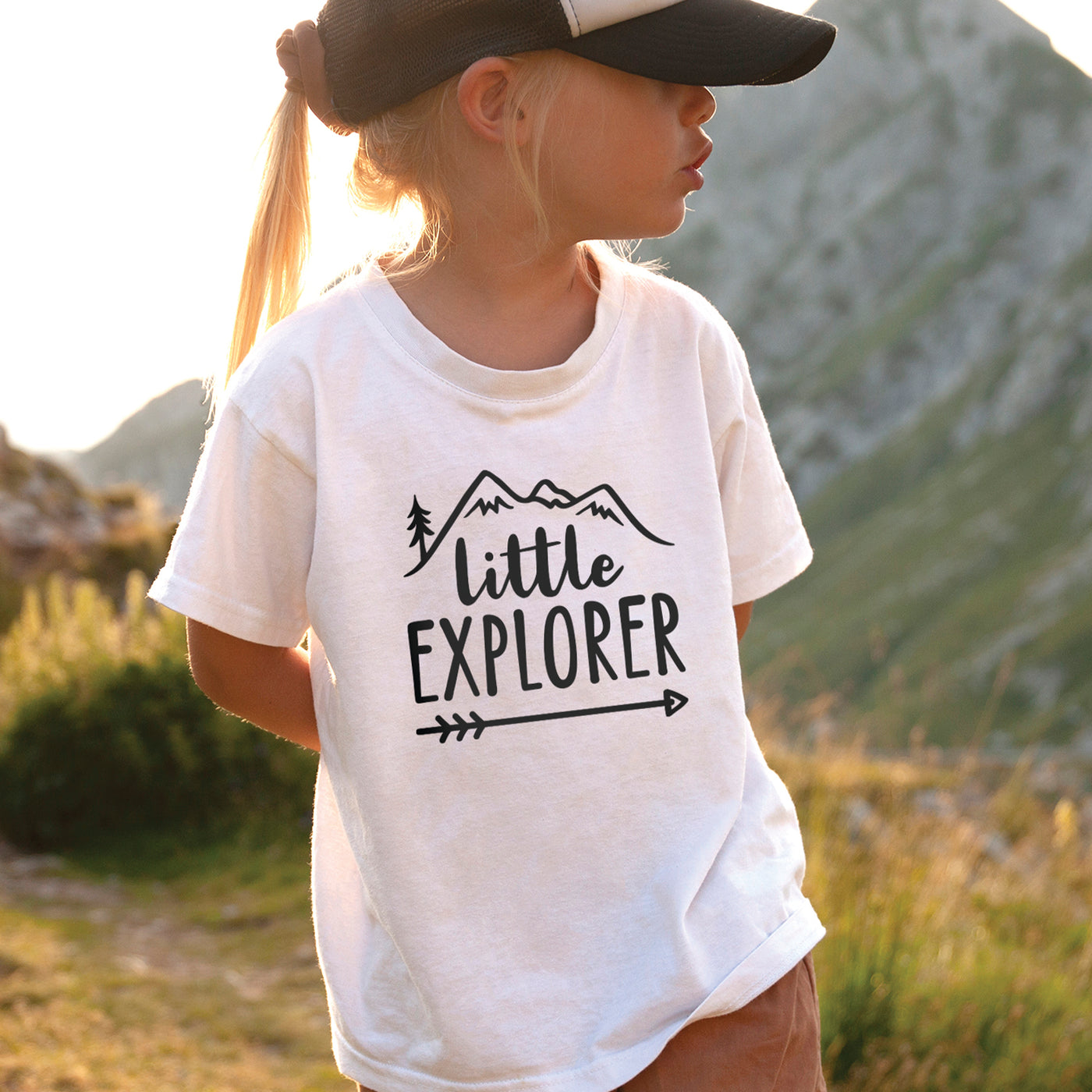 little girl in nature wearing white tshirt with black little explorer text and mountains pine tree and arrow print
