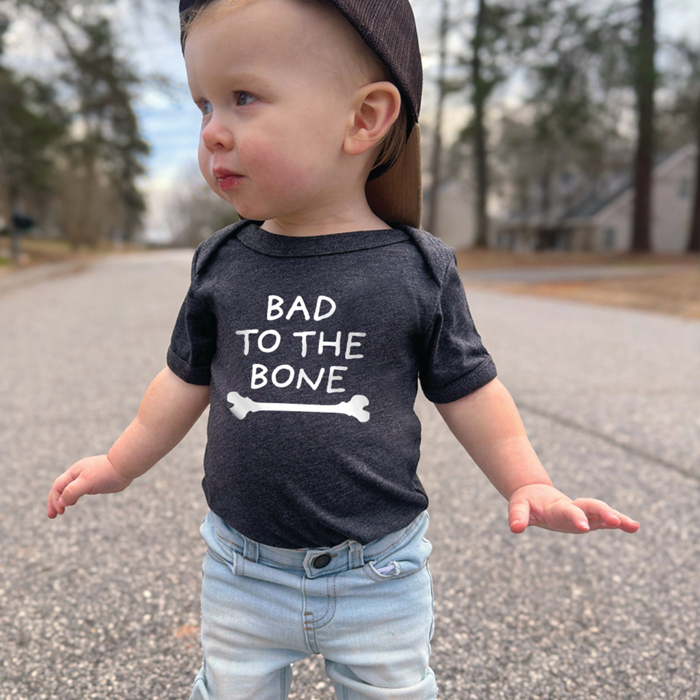 Little boy wearing short sleeve white onesie with black yellow orange chicks dig me print, standing outside wearing a checkered black and white hat and jeans 