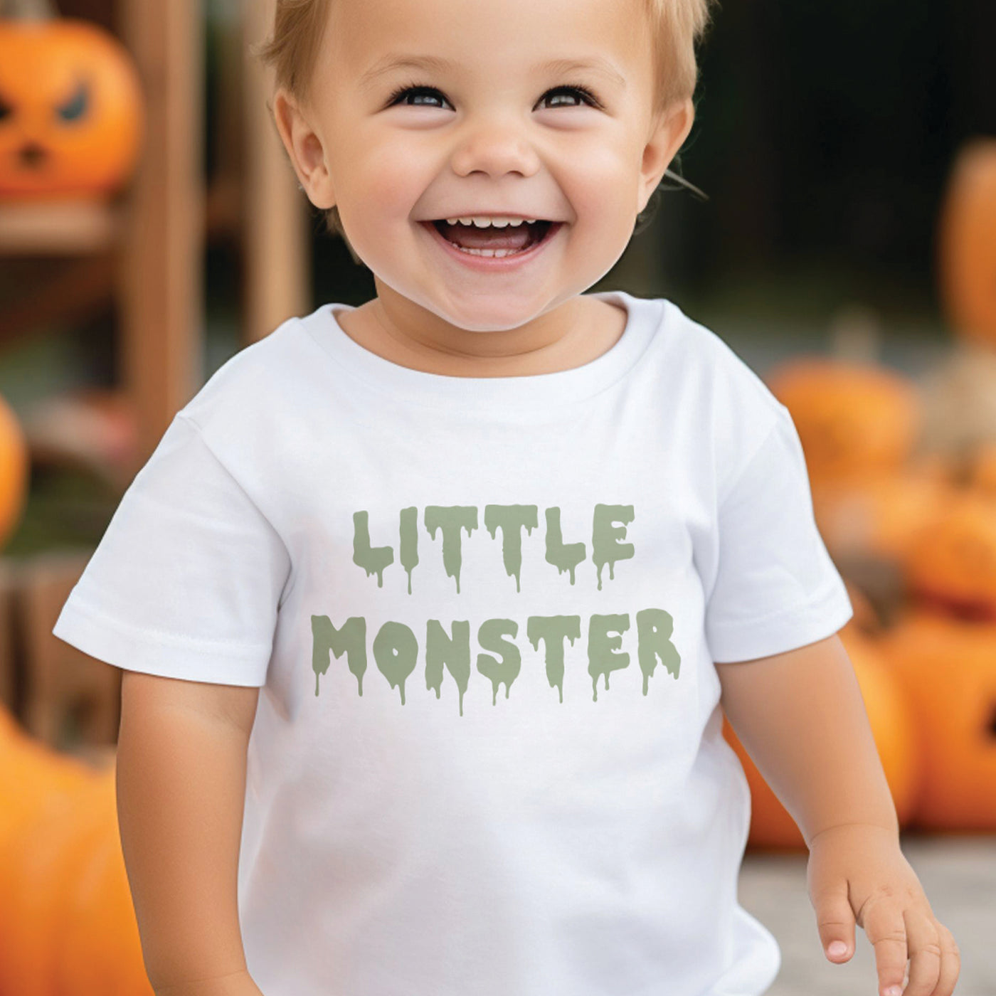 cute little boy wearing white tshirt with sage green little monster dripping text print, standing in front of halloween jack o lantern pumpkins