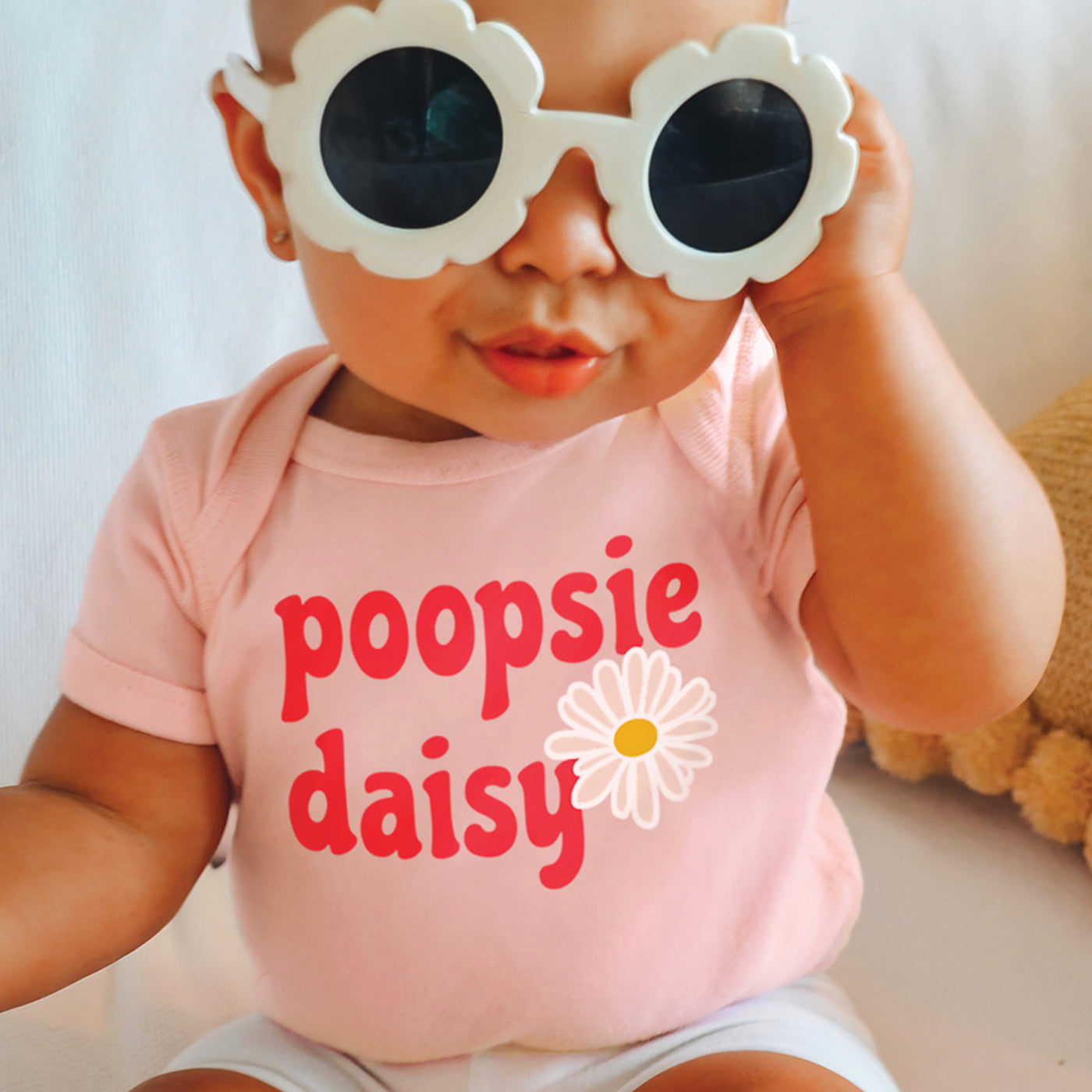 Little baby girl wearing short sleeve peach onesie with pink white and mustard yellow daisy flower and poopsie daisy text print, sitting on couch wearing cute white retro daisy sunglasses and posing 