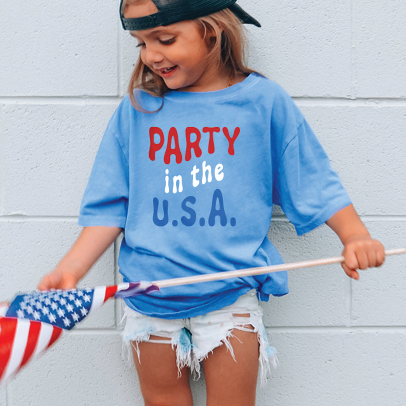 Little girl wearing heather Blue toddler crew neck t-shirt with retro wavy red white and blue party in the USA print , standing outside holding american flag and wearing backwards hat