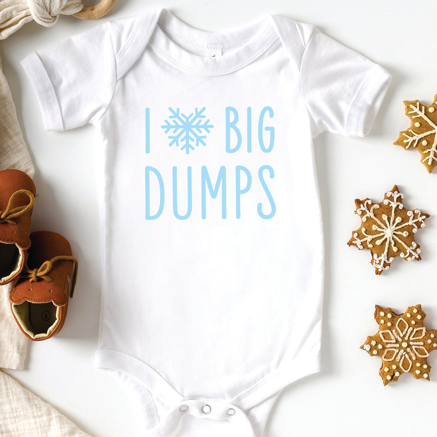 Heather dark grey short sleeve baby onesie with light blue i love big dumps heart shaped snowflake print, laying flat on white background with winter boots, a beanie hat and pine tree branch