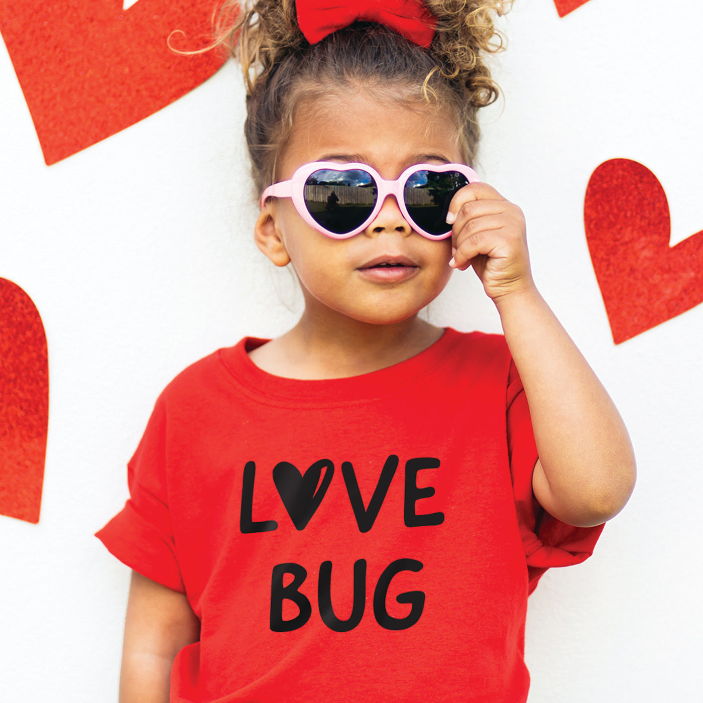 Little toddler girl wearing red short sleeve tshirt with black heart love bug print, standing posing with pink sunglasses in front of dark background