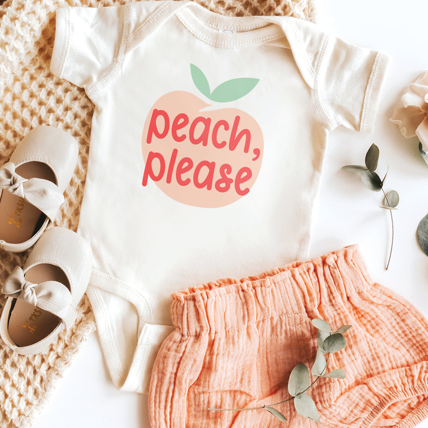 natural white short sleeve baby onesie with peach, pink and green peach please text print inside peach fruit design, laying flat on white background styled with pink shorts shoes and flowers