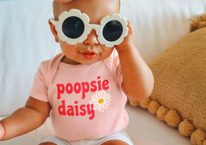 Little infant girl wearing peach one piece bodysuit with coral pink poopsie daisy text with white and mustard yellow daisy flower print, sitting on couch posing with white daisy sunnies