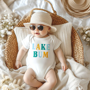 Baby wearing white short sleeve baby bodysuit with alternating light blue, mustard yellow and teal blue LAKE BUM text print on front, laying in wicker basket with blankets and pillow, wearing a sun hat and sunglasses