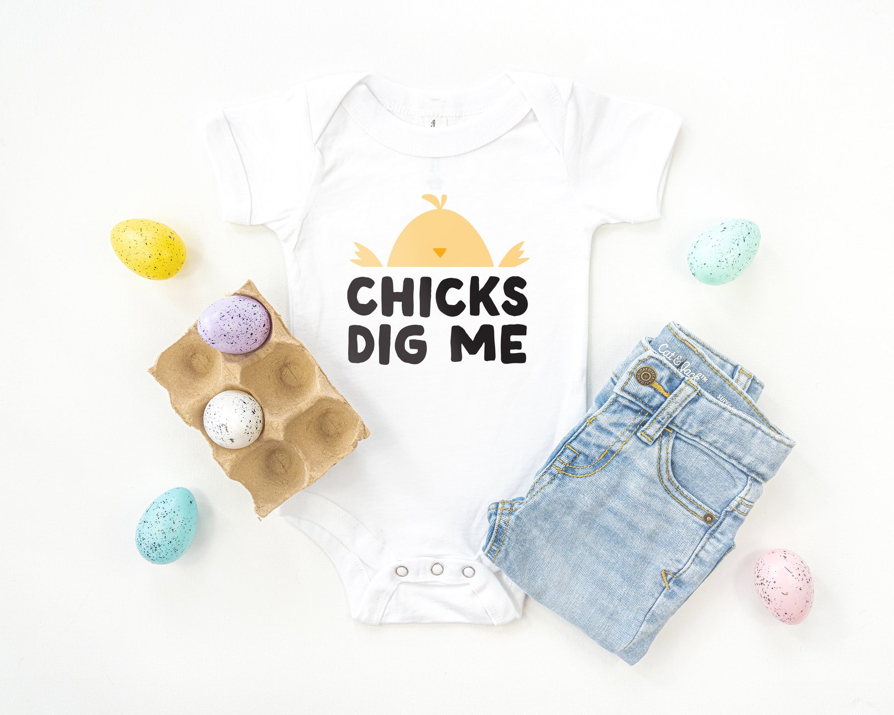 White short sleeve infant bodysuit with yellow, orange and black CHICKS DIG ME with baby chick design printed on the front, laying flat on white background with a pair of jeans and a crate of easter eggs