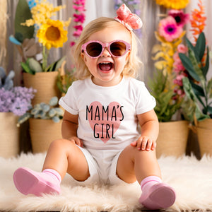Cute little girl wearing white short sleeve toddler shirt with black and pink MAMAS GIRL print inside big heart print, white shorts and pink shoes, sitting on fuzzy blanket in front of flowers smiling and laughing